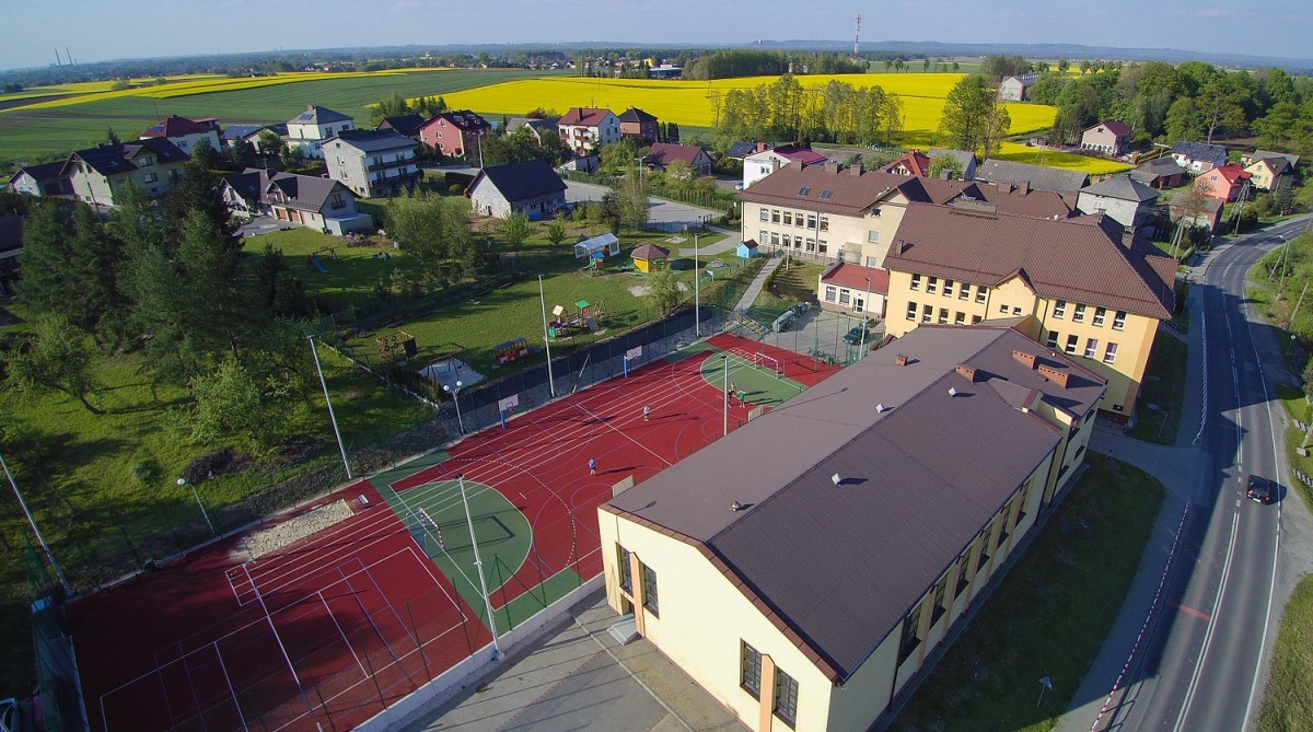 widok z lotu ptaka na boisko sportowe czerwone z białymi liniami, w tle krajobraz pól zółtych i zielonych, dookoła boiska budynki szkoął kremowa z brązowym dachem, oraz budynki mieszkalne