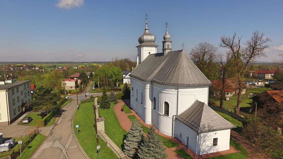 widok na kosciół  biały kościółe z 2 wiezyczkami wokół zabudowania blok oraz domy , dookoła koscioła ogrodzenie, zielona trawa 2 iglaki oraz drzewa. niebieskie niebo - pogodny dzień