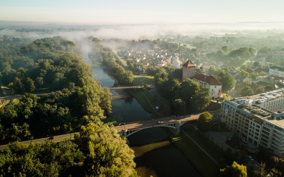 zdjecie  z lotu ptaka na oświęcim po srodkurzeka która przecinają 2 drogi po prawej stronie zamek i zabudowania po lewej str. drzewa w tle krajobraz gór schowany za mgłą