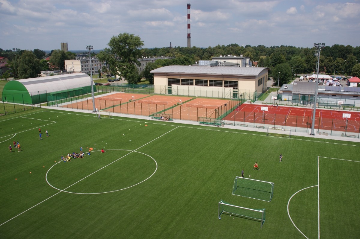 widok z lotu ptaka na centrum sportowe na 1 planie boisko do piłki nożnej z zielona murawa i białymi liniami graja na nimz awodnicy, po lewej stronie kryty kort i 3 bez zadaszenia,a po prawej boisko wielofunkcyjne czerwone z białymi liniami w tle drzewa