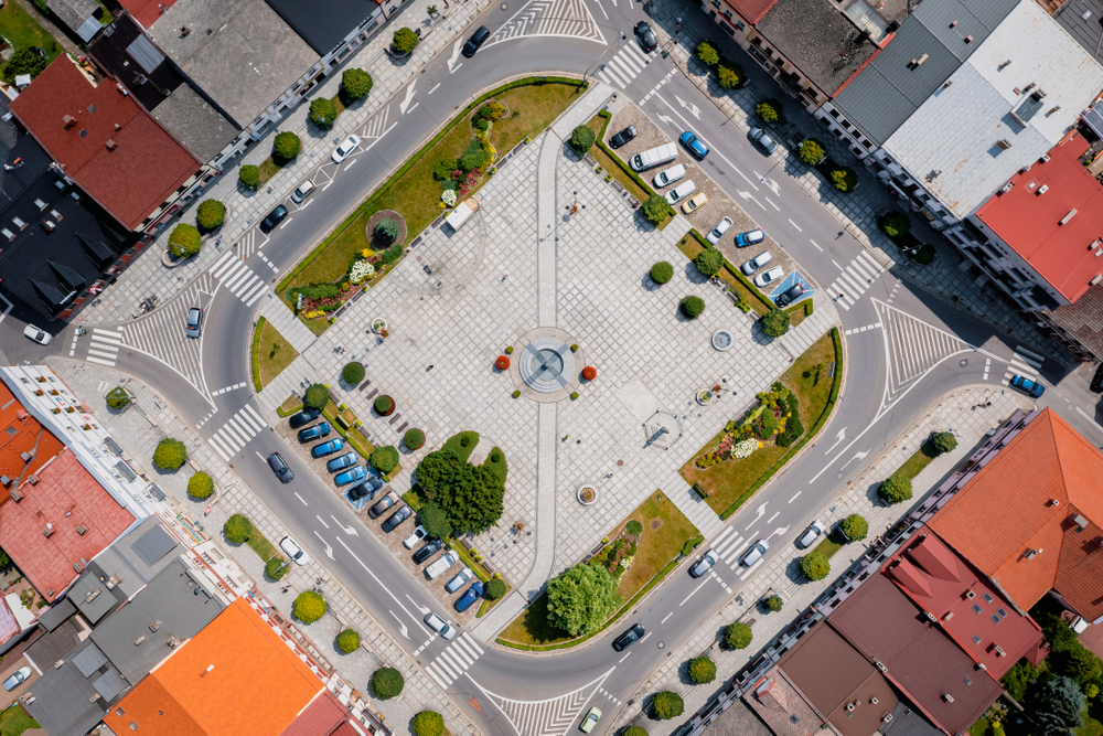 WIDOK Z LOTU PTAKA NA RYNEK PO ŚRODKU RYNEK W FORMIE KWADRATU SZARE KOSTKI DOOKOŁA DACHY BUDYNKÓW W KOLORACH CZERWONYM, RUDYM CZARNYM , PO PEAWEJ I LEWEJ STR PARKINGI ZA AUTAMI, A PO DWÓCH PRZECIWLEGŁYCH ZIELEŃ DRZEWA I KRZEWY