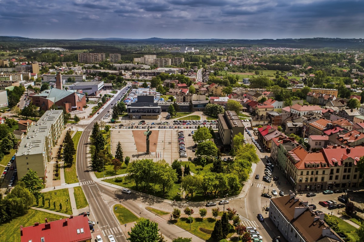 WIDOK NA CHRZANÓW Z LOTU PTAKA POSRODKU PLAC TYSIĄCLECIA Z SZAREJ KOSTKI, PO ŚRODKU POMNIK ORŁA WYSOKI, ORZEŁ W KOLORZE ZIELONKAWO-NIEBIESKIM, DOOKOŁA ZABUDOWANIA KAMIENICE, BLOKI, IINSTYTUCJE KULTURY, W TLE WIDOK NA OSIEDLA Z WIEŻOWCAMI, ZIELEN