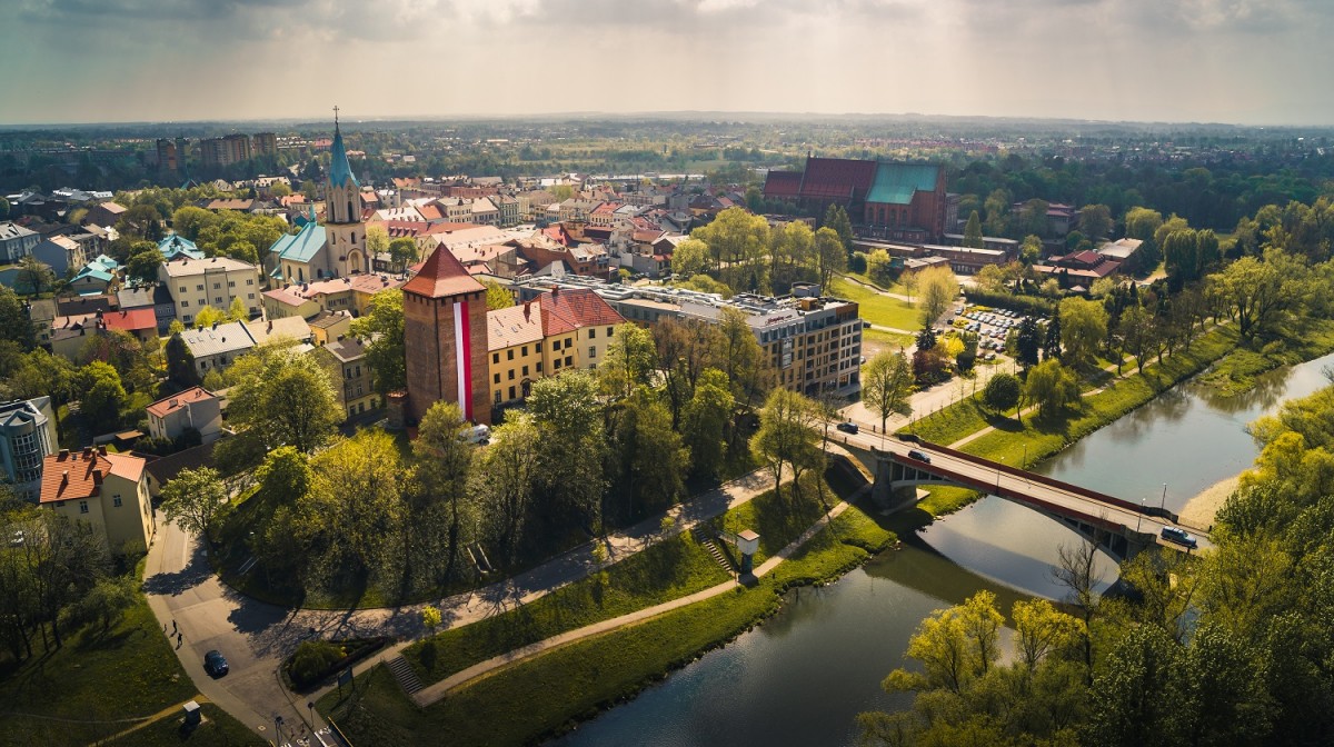 WIDOK NA STARE MIASTO Z LOTU PTAKA PO ŚRODKU RYNEK Z KAMIENICAMI I KOSCIOŁEM PO BOKACH ZABUDOWANIA NA 1 PLANIE RZEKA SOŁA, NA PRZEGACH TRAWA, RZEKĘ PRZECINA MOST 