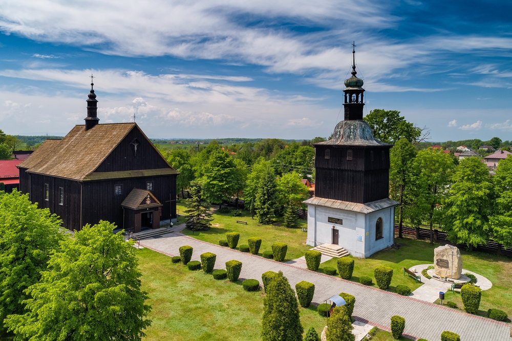 Drewniany zabytkowy kościół z dzwonnicą wśród drzew.