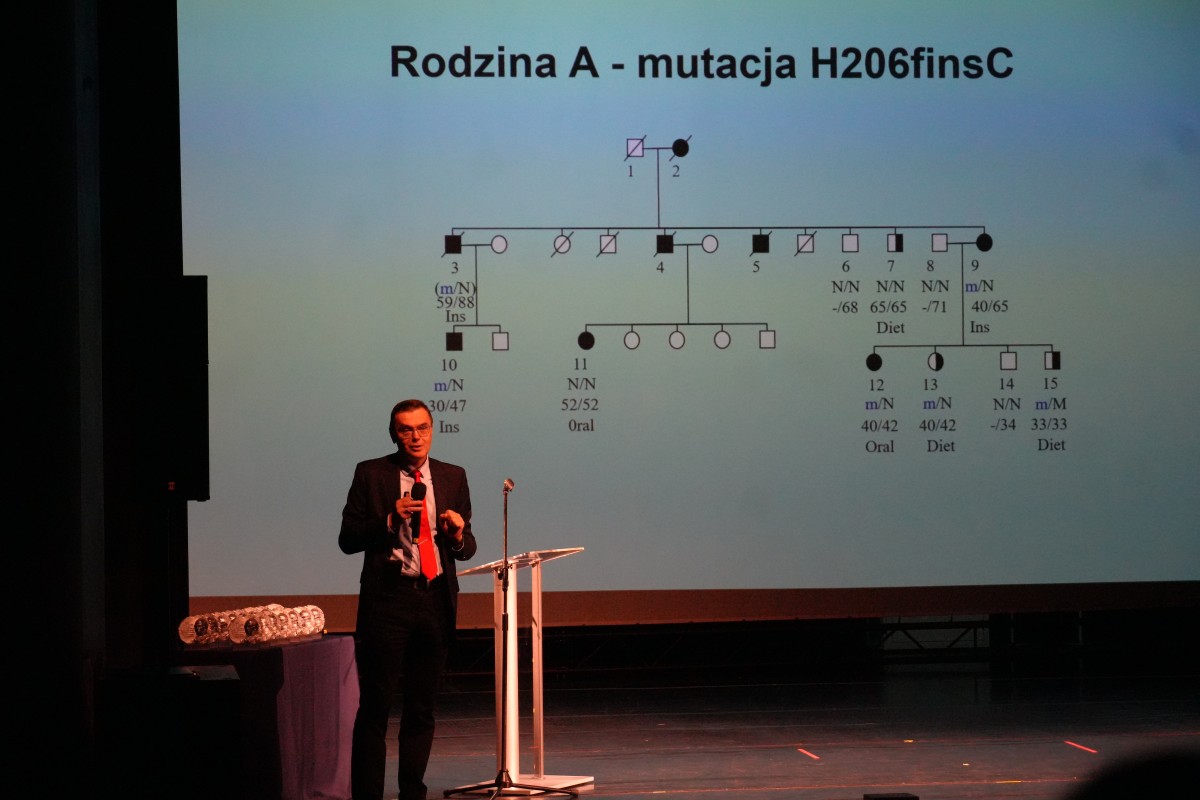 Zdjęcie mężczyzny przemawiajacego zza mównicy. W tle prezentacja z drzewem genealogicznym.