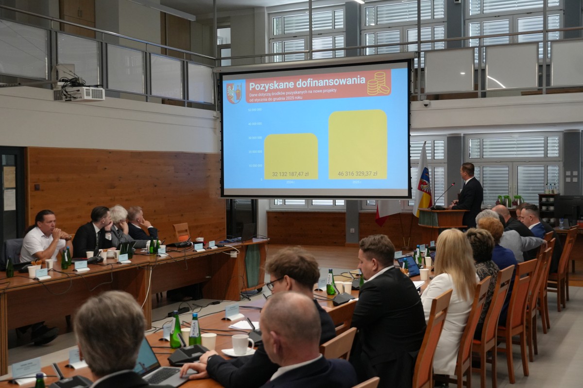 Mężczyzna stoi za mównicom, zerkając na prezentacje przemawia. Pozostałe osoby siedzą przy stołach i patrzą na prezentacje. 