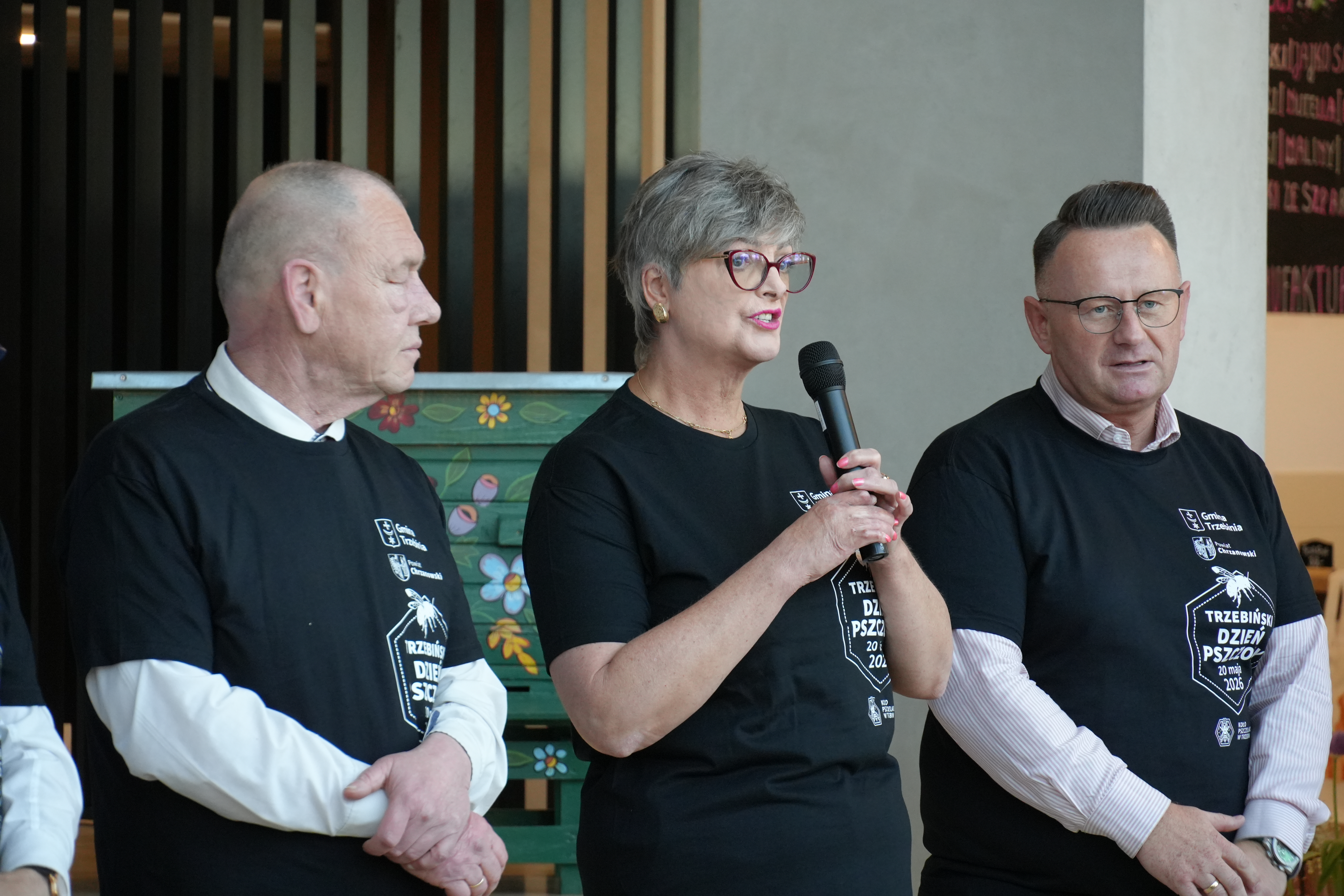 Kobieta oraz dwóch mężczyz stoi obok siebie. Kobieta trzyma mikrofon i przemawia.