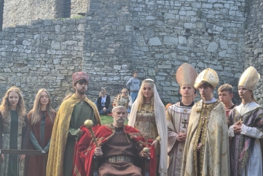 Inscenizacja koronacji: król na tronie, w prawej ręce berło, w lewej jabłko królewskie, obok m.in. duchowieństwo 
