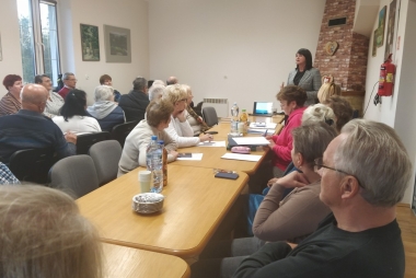  Starsze osoby siedzą przy stole, kobieta przemawia na stojąco 
