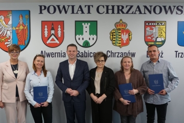 Do zdjęcia pozują: cztery kobiety i dwóch mężczyzn. Część z nich trzyma w ręku teczki. W tle herby