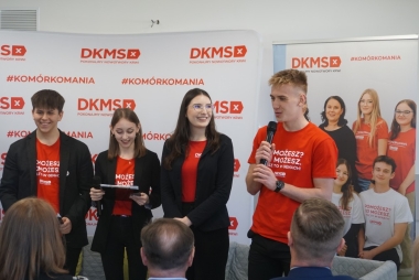 Zdjęcie 4 młodych osób. Za nimi roll'upy i ścianki promocyjne. Jeden z uczniów trzyma mikrofon i przemawa do grupy osób.