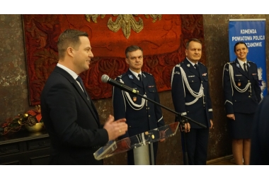 Zdjęcie czterech osób. Trzy osoby stoją w mundurach, jedna w graniturze za mównicą.