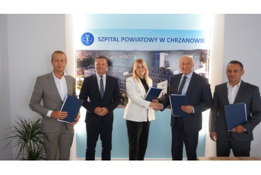 Czterech mężczyzn w garniturach i kobieta w białym żakiecie. W tle zdjęcie szpitala 