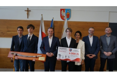 Zdjęcie w sali z flagami oraz herbami stoi obok siebie siedem osób. Dwójka z nich trzyma szablę oraz kartonowy voucher.